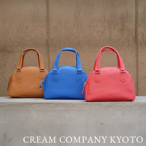 三款牛皮半月迷你手提包，分別為淺棕色、亮藍色及珊瑚紅色，皆帶有細緻的皮革紋理和圓潤的提把設計。