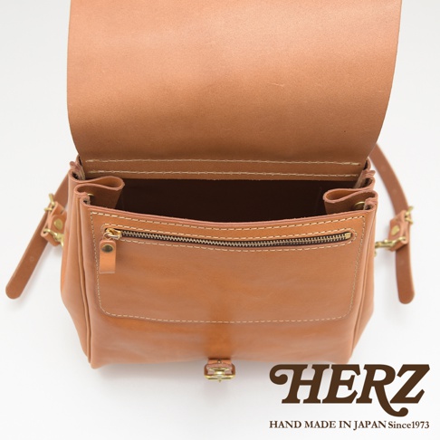 焦糖色皮革後背包，帶有拉鍊前袋和金屬釦，內部空間可見，展現職人手作質感。