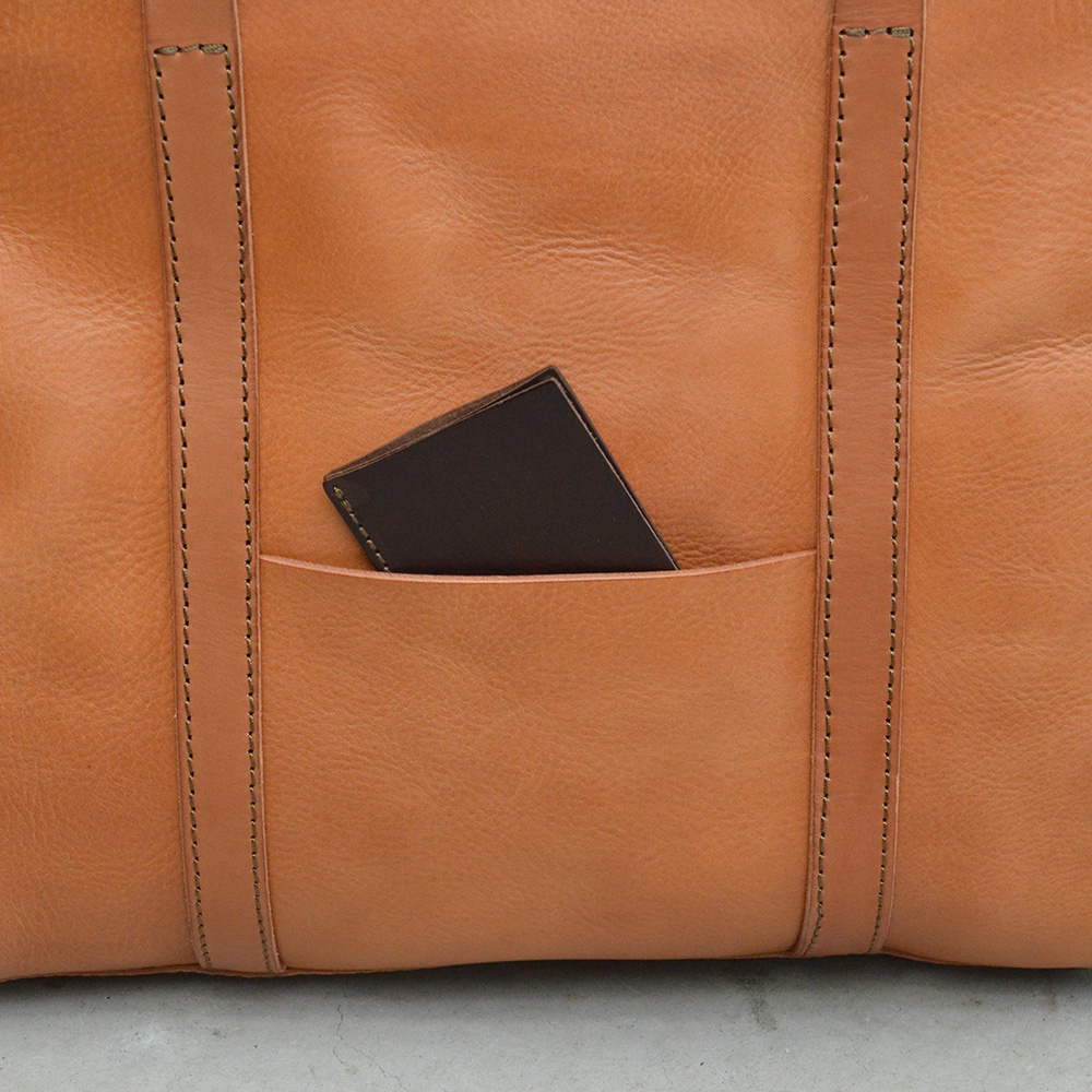 棕褐色皮革托特包，有外側口袋，口袋裡放著一張深棕色卡片。包包表面有縫線裝飾，材質呈現自然紋理。
