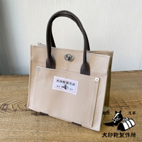 淺卡其色純棉帆布迷你托特包，配有深棕色提把和銀色旋轉鎖扣。包包前方有一個大口袋，上面貼有印有日文的白色標籤，標籤上有狗和袋子的圖案。