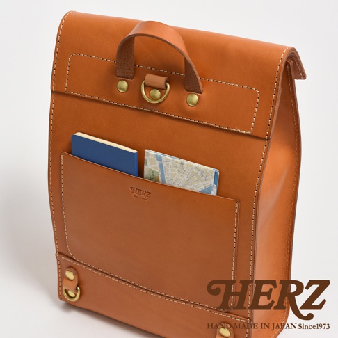 職人手作皮革紙袋風後背包，呈現自然的棕褐色，材質為皮革，設計簡約，前方有一個大型外袋，內有藍色筆記本和地圖。