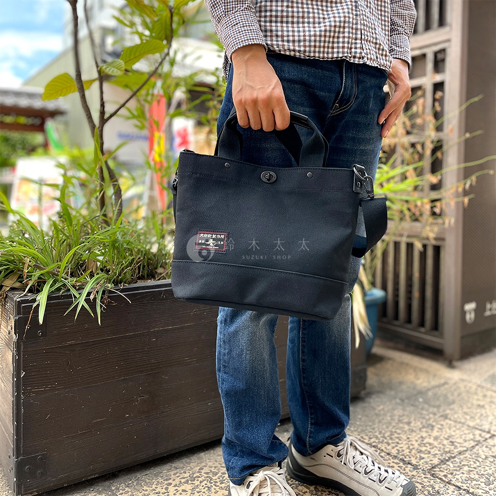 黑色帆布材質的兩用托特包，包身正面有標籤，配有提把和可拆卸肩帶。