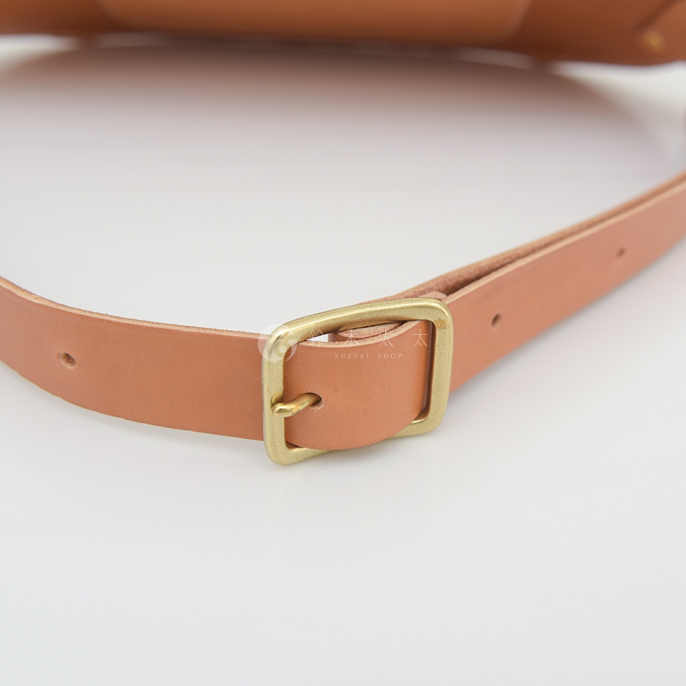 一塊淺棕色皮革的斜背包，上面有一個黃銅色帶扣，皮帶上有調整孔，扣子旁有 SUZUKI SHOP 字樣