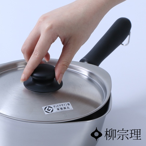 一隻手輕輕掀開柳宗理不鏽鋼霧面單手鍋的鍋蓋，鍋蓋上貼有象徵優良設計的標籤，整體呈現金屬質感與簡約工業設計風格，右下角印有柳宗理品牌名稱。