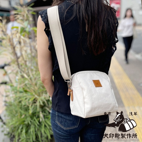 一位女士身穿黑色上衣和牛仔褲，側背一個白色石洗帆布散步包，包包上有一個皮革標籤，旁有東京淺草犬印製作所的圖案。