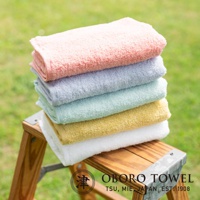 堆疊的五條毛巾，顏色由上至下分別為粉紅色、淺藍色、淺綠色、鵝黃色和白色。毛巾質地柔軟，表面呈現細緻的紋理，擺放在木製架子上，背景是綠色的草地。