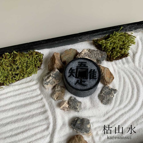 日本枯山水模型，白色細砂鋪陳，有著細膩的紋路，散落著深淺不一的灰色石頭。中央擺放一枚黑色圓形裝飾，上面刻有漢字。兩側點綴著翠綠的苔蘚，營造出簡約禪意。