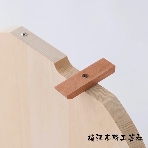 木製圓形砧板，淺木色，表面有木紋，一側有銀色金屬釘固定一個深棕色木塊，木塊上有螺絲。