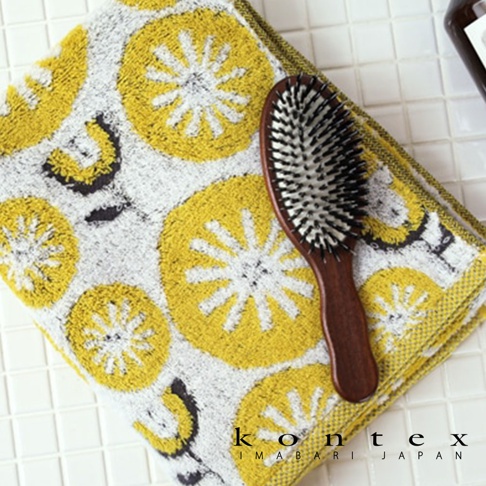 模式B 白色毛巾上印有黃色檸檬花圖案，一旁的棕色木柄髮刷放在毛巾上，毛巾右下角印有 KONTEX IMABARI JAPAN 字樣。