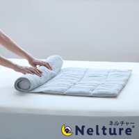 一隻手正在將一條淺灰色的毛巾枕捲起，毛巾枕鋪在白色的床墊上，枕頭上有縫線分隔的格子，右下角有 Nelture 品牌標誌，標誌為一彎月亮上有隻綿羊，旁邊是日文「ネルチャー」及英文「Nelture」。