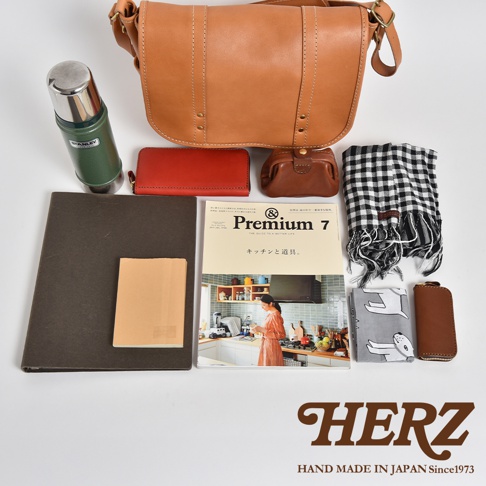 職人手作皮革復古郵差包，搭配一個綠色保溫瓶、一個紅色皮夾、一個棕色零錢包、一本雜誌、一條格紋圍巾、一個筆記本及一個鑰匙包。