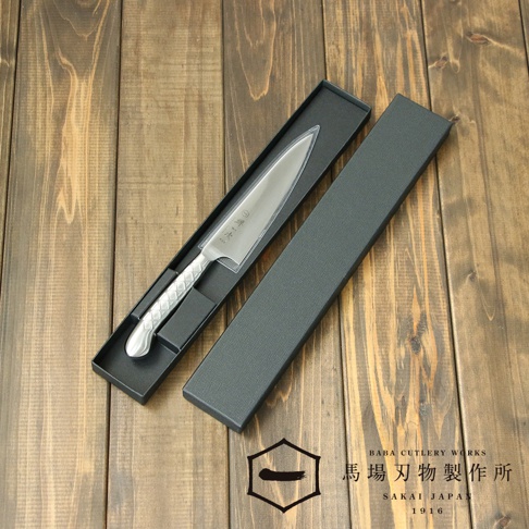 [堺一次] 不鏽鋼水果刀，刀刃呈現銀色金屬光澤，握柄為銀色不鏽鋼材質，帶有菱形格紋，收納於黑色長形紙盒內，背景為木紋桌面。