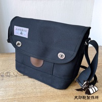 深藍色純綿帆布製成的郵差包，帶有兩個銀色金屬扣和一個棕色皮革裝飾。包的左上方有一個帶有日文文字和狗圖案的標籤。