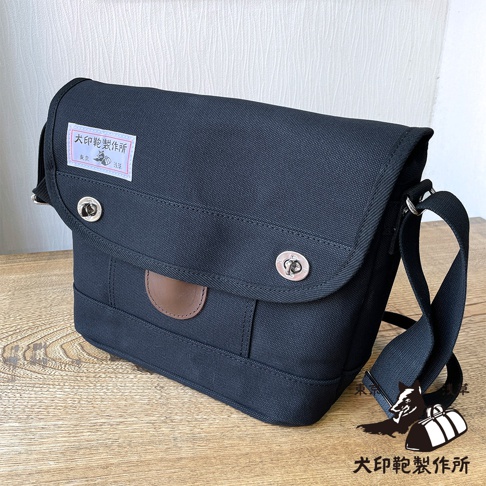 深藍色純綿帆布製成的郵差包，帶有兩個銀色金屬扣和一個棕色皮革裝飾。包的左上方有一個帶有日文文字和狗圖案的標籤。