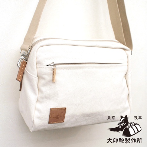 一個米白色帆布斜背包，正面有拉鍊口袋，右下角有一個棕色皮革標籤，上面有圖案和文字，旁邊還有東京淺草犬印鞄製作所的圖案和文字。