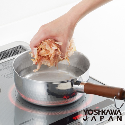 鄉技不鏽鋼單柄雪平鍋，鍋身為銀色不鏽鋼材質，帶有錘紋，配有木質手柄。鍋中裝有水，一隻手正將柴魚片放入鍋中。鍋子放置於電磁爐上，電磁爐發出紅光。