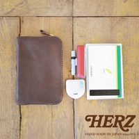 職人手作皮革Organ雙拉鍊收納袋，深棕色荔枝紋皮革，附有黃銅拉鍊與皮革拉環。旁邊擺放著一支白色筆、紅色印章盒、uni白色小物及一本綠色邊條的存摺。