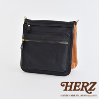 黑色皮革翻轉雙色直式斜背包，設有金色拉鍊前袋及頂部拉鍊開口，搭配棕色皮革內襯。