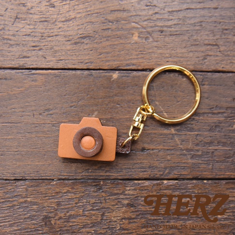 職人手作皮革相機造型鑰匙圈，主體為淺棕色皮革，鏡頭部分為深棕色皮革點綴，搭配金色金屬鑰匙圈，整體精緻可愛。