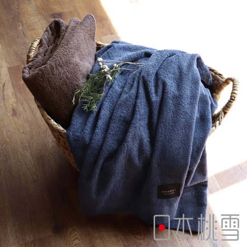 編織籃中擺放著一深棕色與一深藍色的超長棉毛巾被，質地柔軟，深藍色毛巾被上裝飾有幾枝松枝，毛巾被的標籤清晰可見，顯示品牌名稱。
