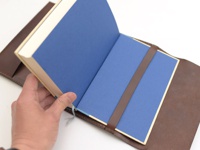 職人手作皮革翻蓋記事本書套，深棕色皮革材質，內頁為藍色。書套內有皮革束帶固定書本。