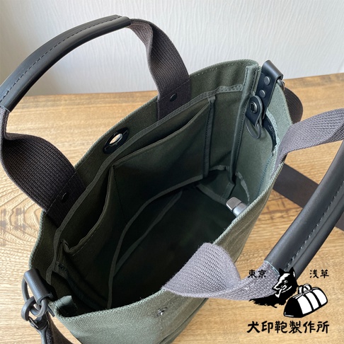 軍綠色帆布製成的兩用托特包，設有黑色皮革提把與肩帶，包內有多個分隔口袋，設計簡潔實用。