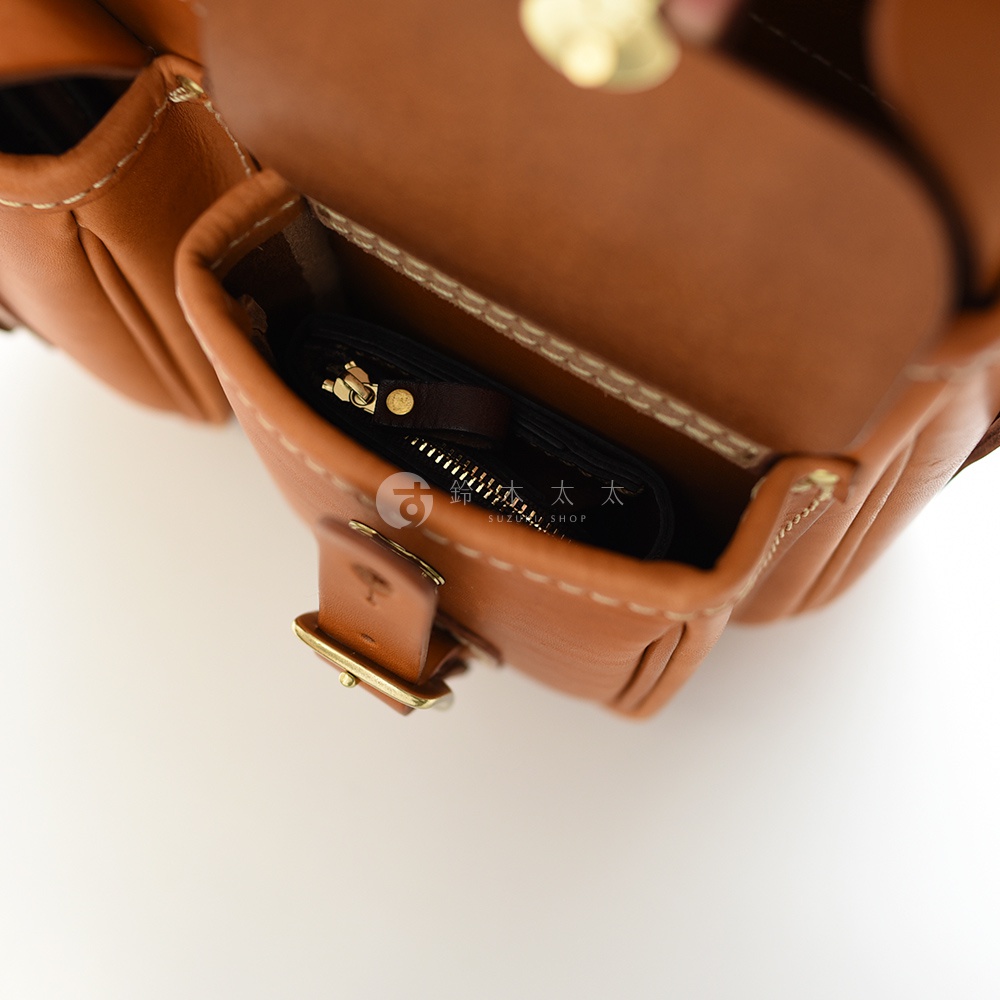 內視圖，展示一個棕色皮革郵差包打開的內部，裡面有一個黑色拉鍊小錢包，上面有金屬拉鍊和一個小棕色皮革吊牌。