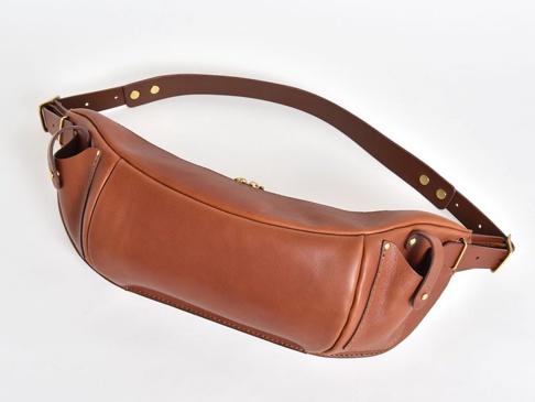 咖啡色皮革製成的大容量斜背包，有可調節的寬背帶，搭配金色金屬扣環和鉚釘，包身線條流暢，質感溫潤。
