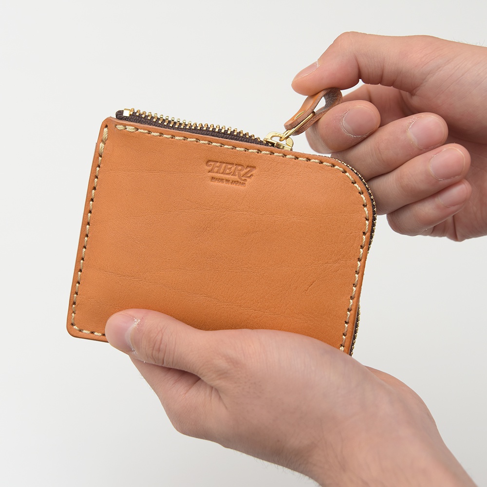 職人手作皮革L型拉鍊迷你短夾，皮革顏色為棕褐色，飾有品牌燙金Logo，搭配黃銅色拉鍊及縫線，由雙手持握展示。