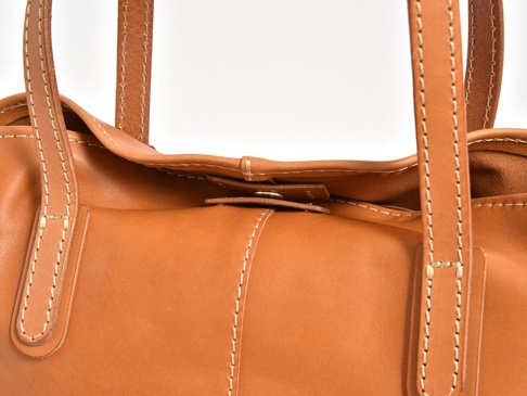 職人手作皮革橫式肩背托特包，採用溫暖的棕褐色皮革，搭配奶白色的縫線，展現細緻的工藝。包包的皮革表面光滑，部分細節處可見自然的紋理。