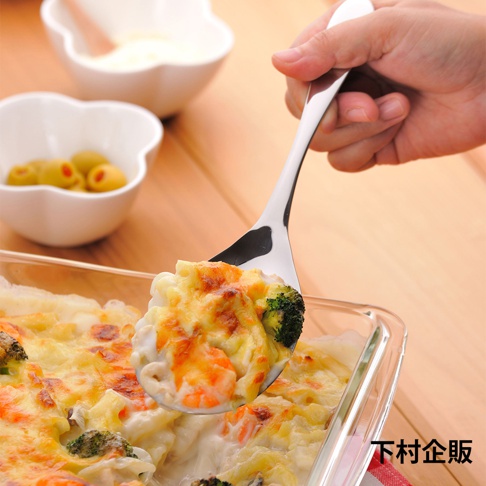 手持不鏽鋼料理分食匙，舀起裝滿焗烤料理的玻璃烤盤，料理中可見花椰菜、蝦仁與起司。