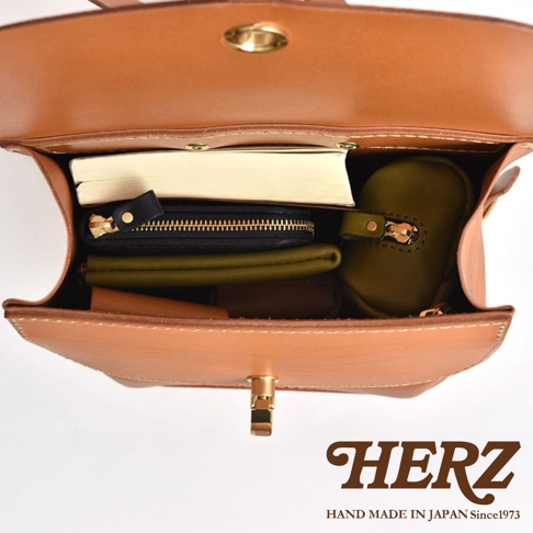 職人手作皮革翻蓋迷你後背包，淺棕色皮革，內部收納物包含書本、黑色拉鍊長夾、綠色零錢包及其他小物，金屬鎖扣。