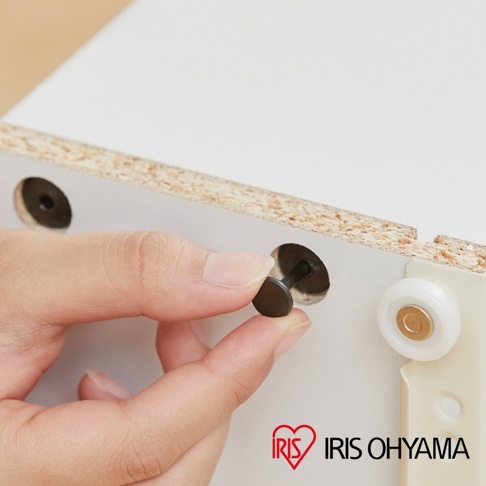 特寫鏡頭顯示一隻手正將一個黑色圓形塑膠扣件安裝至白色木製板材的圓孔中，旁邊安裝有一枚白色滾輪，右下角印有品牌名稱 IRIS OHYAMA。