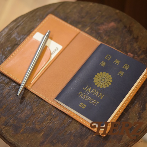 職人手作皮革經典護照套，淺棕色皮革，內含一本深藍色日本護照和一支銀色金屬筆，置於木質桌面。