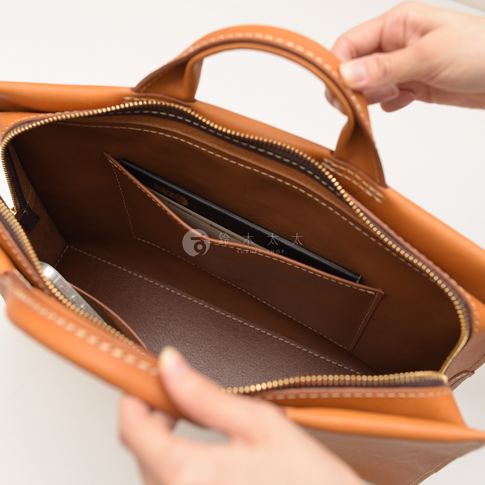 皮革公事包內部，可見手機、卡夾和一本筆記本，提把由手工縫製，品牌標誌印在內襯。