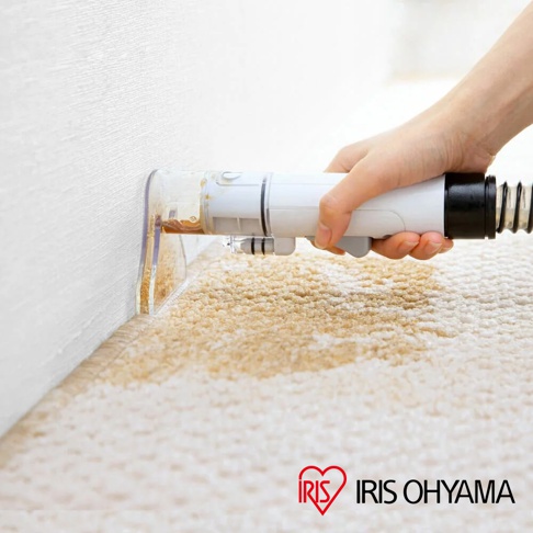 手持白色自動給水織物清潔機，清潔機頭的透明部分正對著米白色絨毛地毯的污漬，機身有黑色軟管連接。