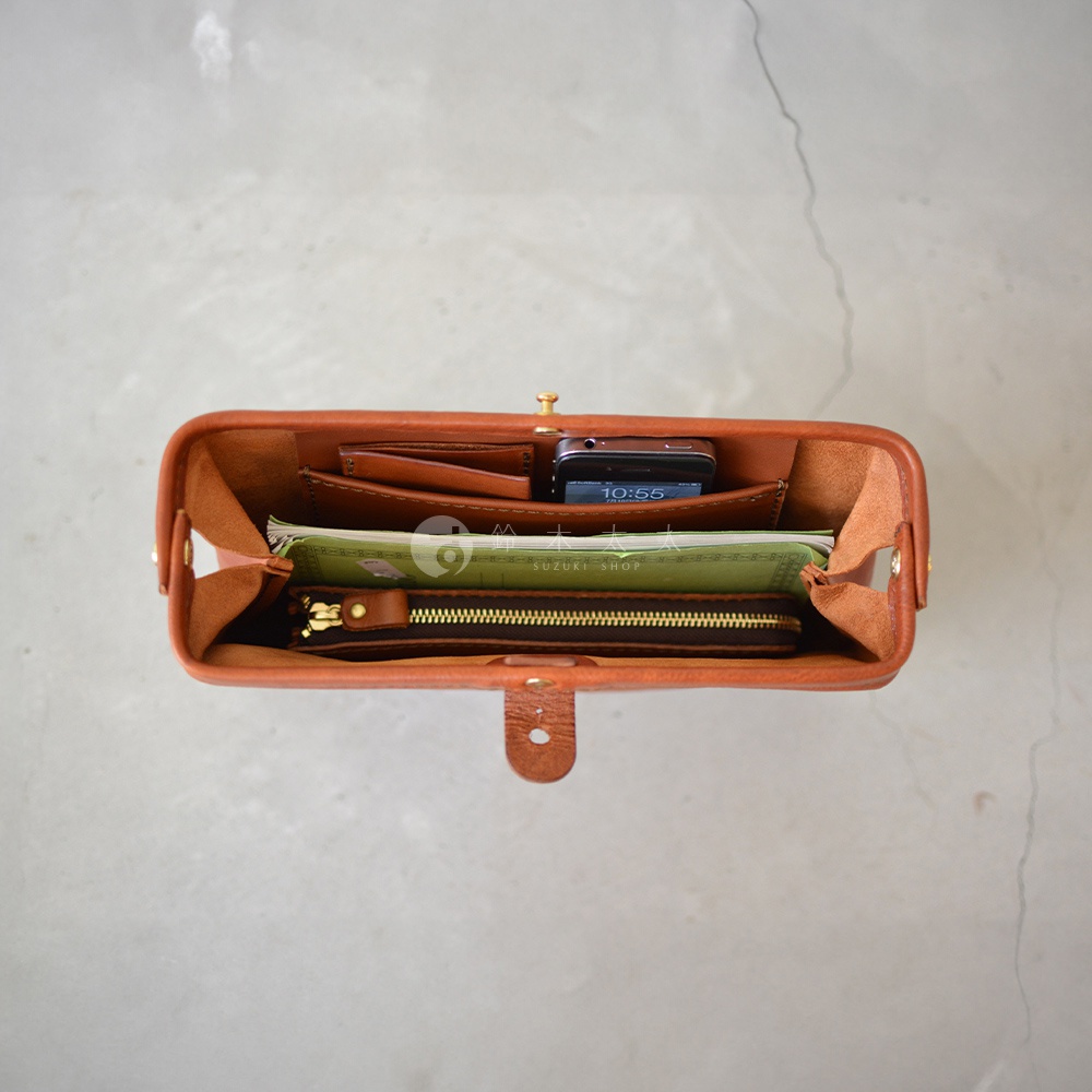 職人手作皮革Organ手拿包，焦糖棕色，展示包內裝有手機、鈔票、卡片夾及拉鍊式零錢包，皮革質地，頂部有金色旋鈕式開關及皮革搭扣。