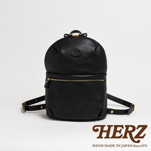 黑色皮革圓角大開口後背包，附有金色拉鍊和肩帶扣環，包包中央有一個品牌壓紋圓標。