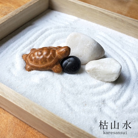 日本枯山水模型，包含一個陶瓷褐色小烏龜擺飾、一顆黑色圓形石子和兩顆白色卵石，整體置於白色細砂上，砂面有耙出的紋路，裝於木框內。