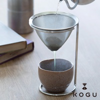 日製18-8不鏽鋼濾茶器，圓錐形細網濾網，搭配銀色金屬立架，正將茶湯濾入下方陶製茶杯中，背景有書本與金屬壺。