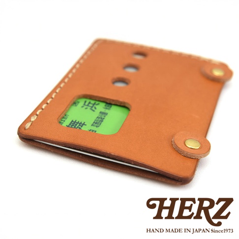 手工製作的棕色皮革卡夾，內部露出一張綠色卡片，上方印有日文文字，品牌標誌HERZ及Hand Made in Japan Since 1973。