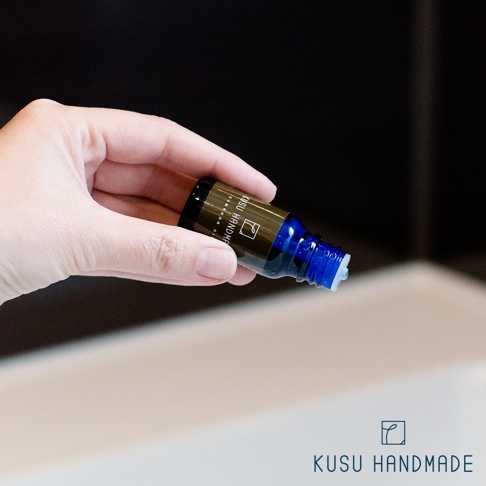 手持一瓶樟楠木條精油，藍色玻璃瓶身，金色標籤印有「KISU HANDM」字樣，瓶蓋處有白色滴管。