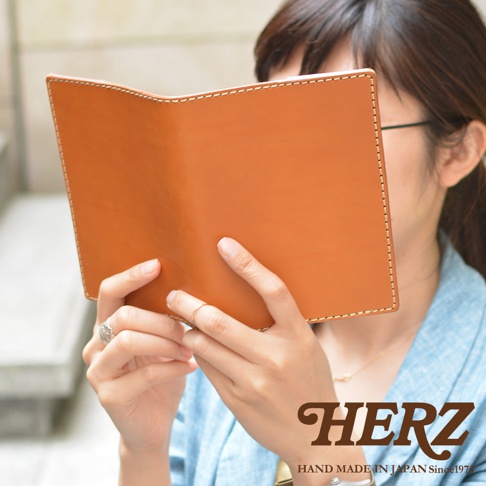 一位女性正在閱讀一本皮革製作的書本，她雙手托著這本棕色皮革書套，皮革表面具有自然的皮革紋理與精緻的縫線工藝，畫面下方標示著日本職人品牌 HERZ 與手作字樣。