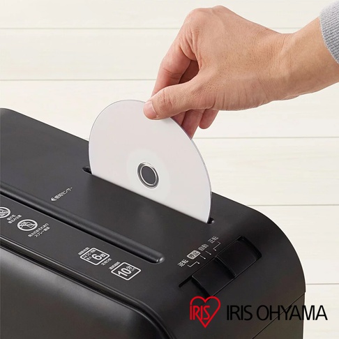 手正將一張白色光碟放入黑色碎紙機的進紙口。碎紙機表面有日文標示和圖示，包括「感應センサー」、「連續碎紙6分鐘」、「過熱保護10分鐘」等，以及「前进」、「停止」、「自動」的切換開關。右下角有「IRIS OHYAMA」的紅色心形標誌。