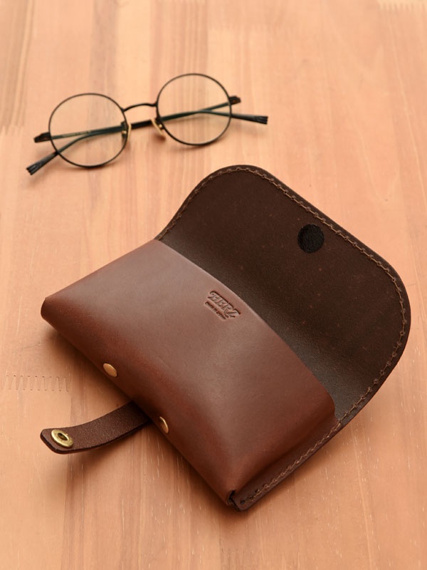 深棕色皮革製成的手工眼鏡收納盒，附有金屬釦帶及內襯，旁邊放著一副圓框眼鏡。