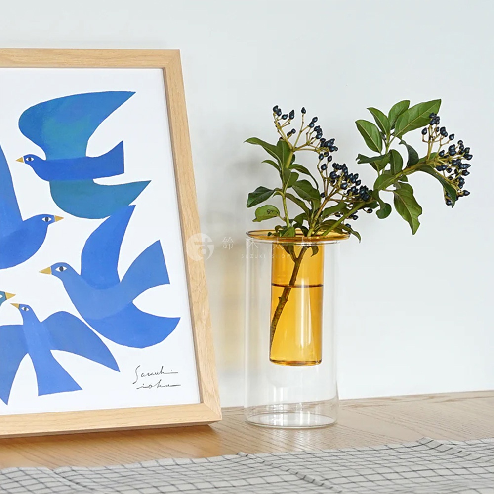 AXCIS PINO 雙層可拆玻璃花瓶-高款，展示於木框畫作旁，花瓶內插有綠葉枝條，瓶身琥珀色玻璃內襯透明外層。