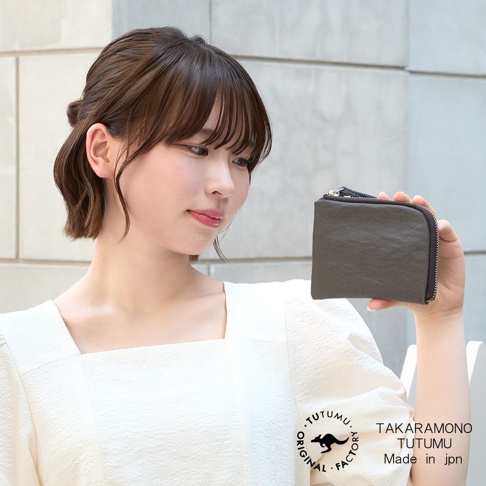 模式B： 一位年輕女子手持一個深棕色迷你拉鍊錢包，臉部側向畫面右方，她身穿一件白色方形領口上衣，圖片右下角有TUTUMU和TAKARAMONO Made in jpn的文字標誌。
