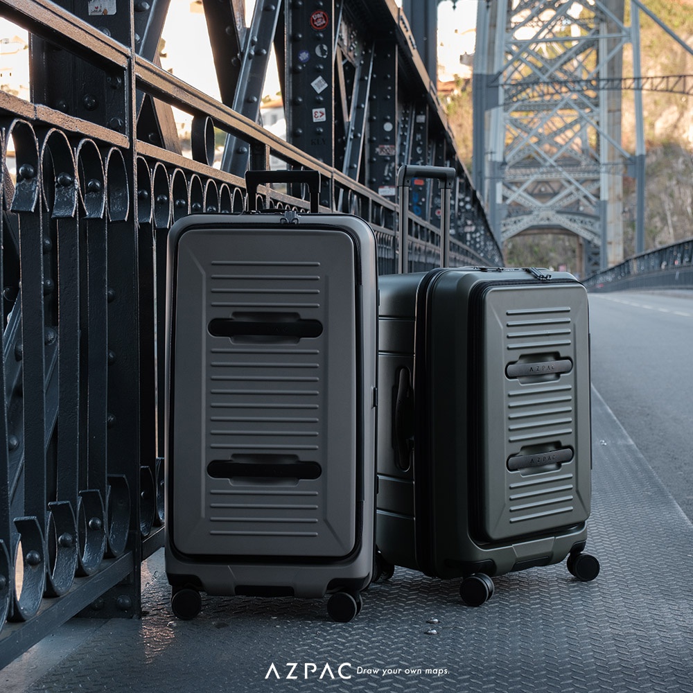 兩款不同顏色的 AZPAC 行李箱放置在鐵橋步道上，箱體表面具有橫向條紋與前開口設計，右側的行李箱拉桿處於升起狀態，整體呈現出戶外旅行的氛圍。