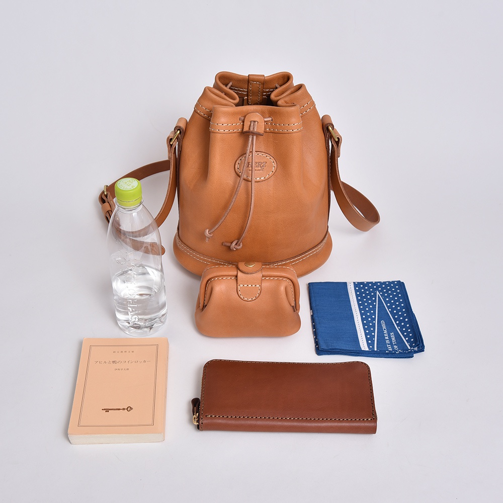 淺棕色皮革束口單肩包，搭配同色系零錢包、長夾、藍色印花手帕和一本淺棕色書籍，以及一瓶礦泉水。