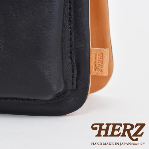 職人手作皮革翻轉雙色直式斜背包，黑色皮革與原色皮革拼接，有壓印品牌標誌的皮革小標籤。